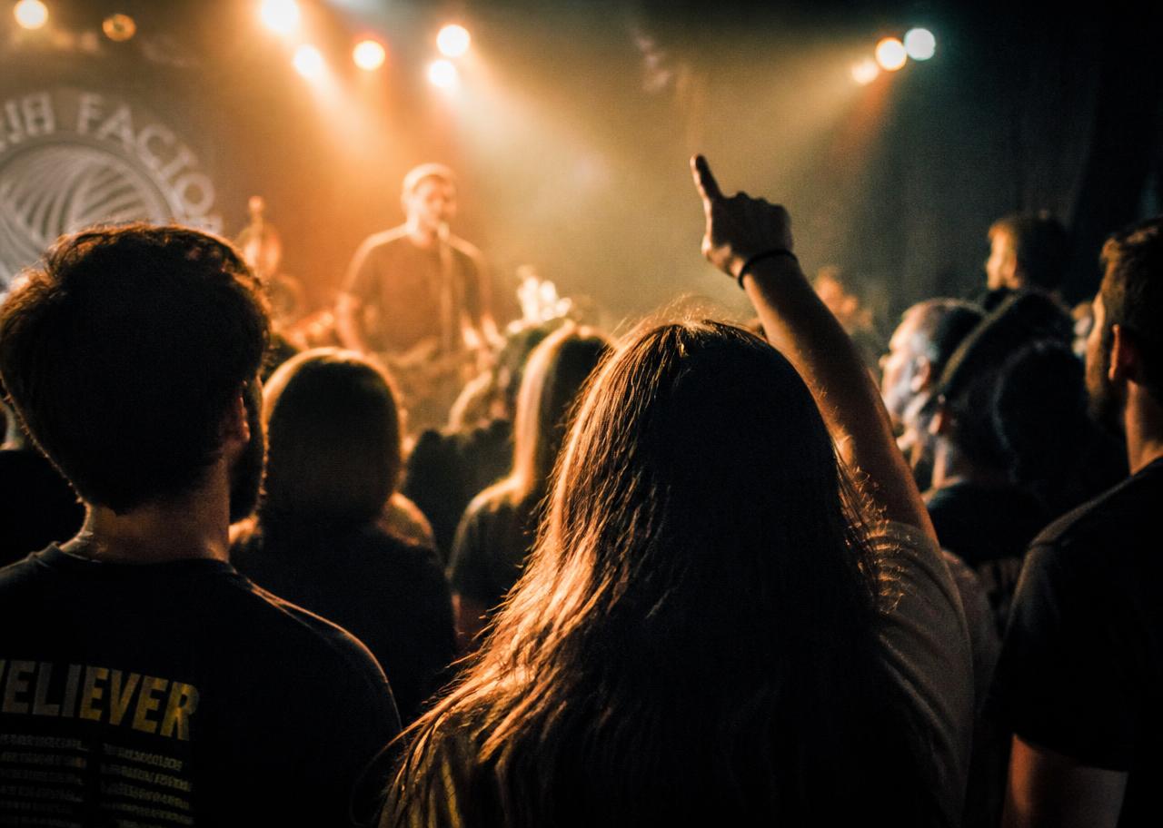 Fans watching an artist from the middle of a small gig crowd, warm stage light ahead